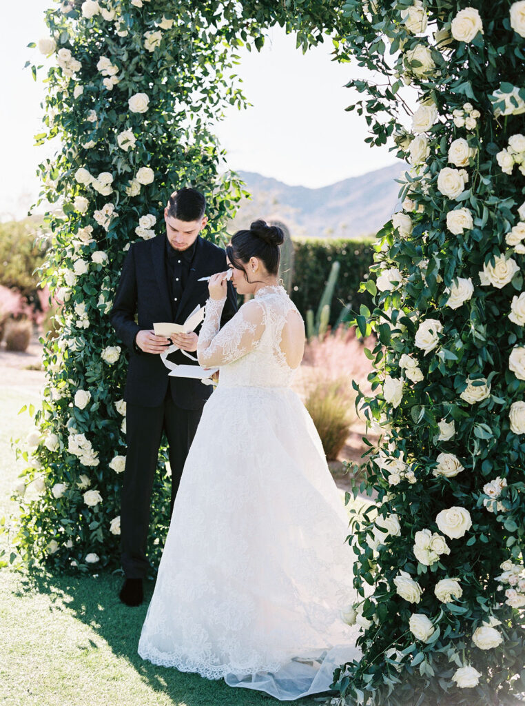 Bride reading vows to groom under floral ceremony arch.