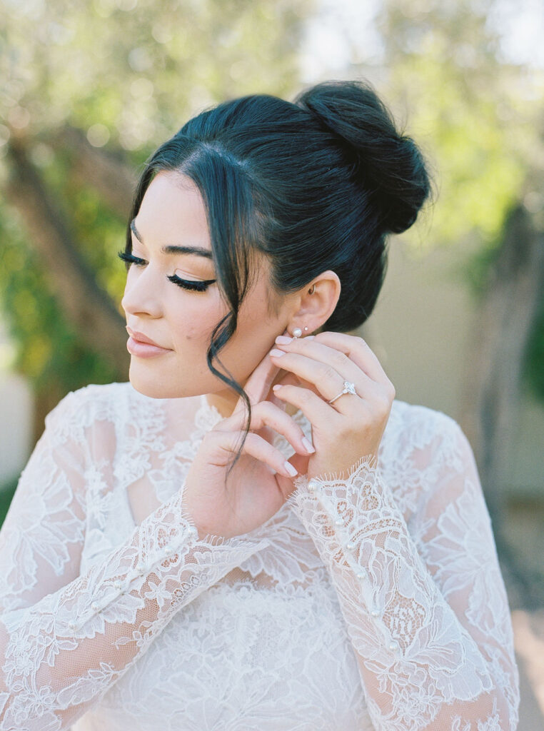 Bride adjusting her earring in lace gown before ceremony.