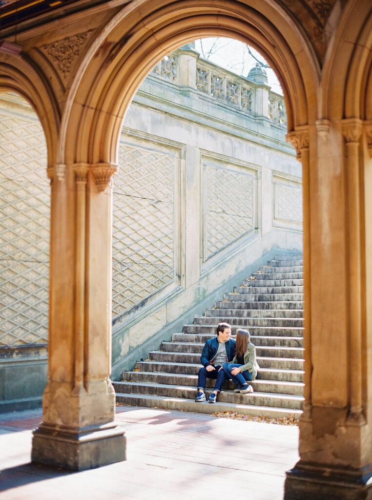 Couple sitting on steps under Bethesda Terrace arch during fall engagement in NYC