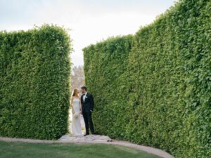 Bride and groom walk through lush green hedge path.