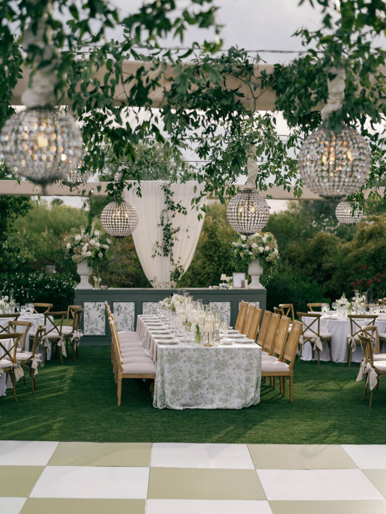 Outdoor wedding reception setup with greenery and chandeliers.