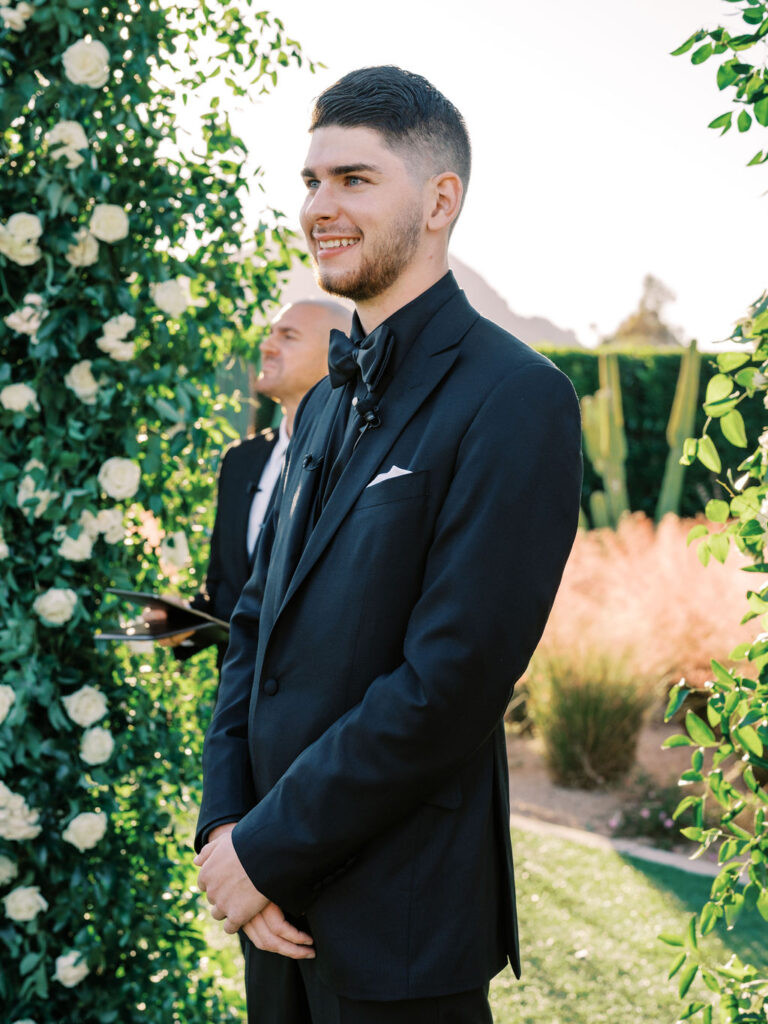 Groom smiling during ceremony under floral wedding arch.