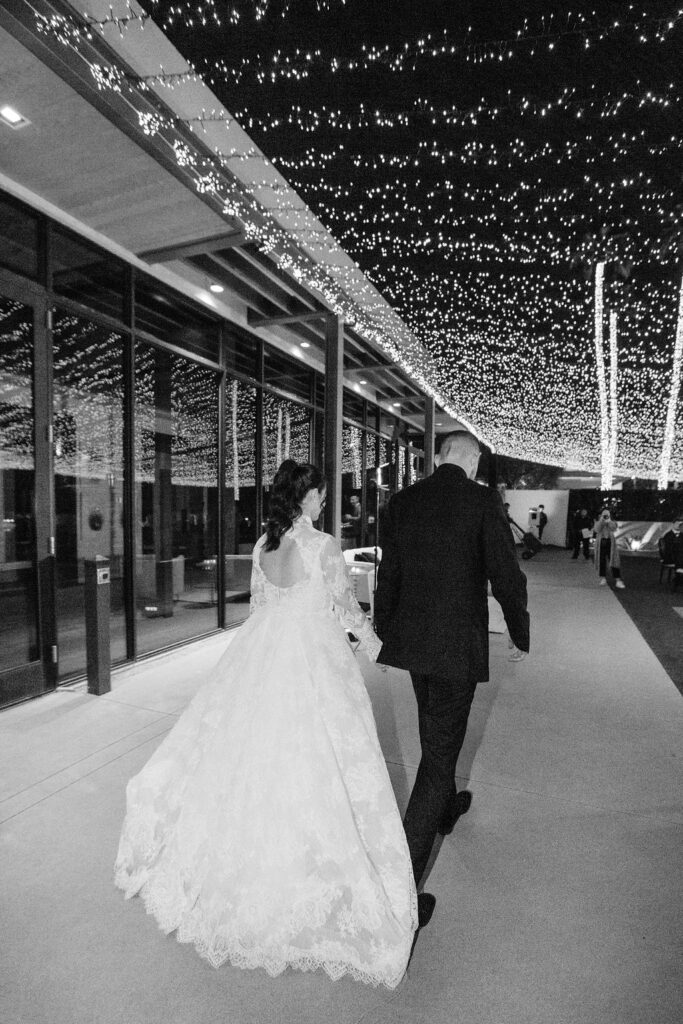 Bride and groom walking hand in hand under glowing string lights.