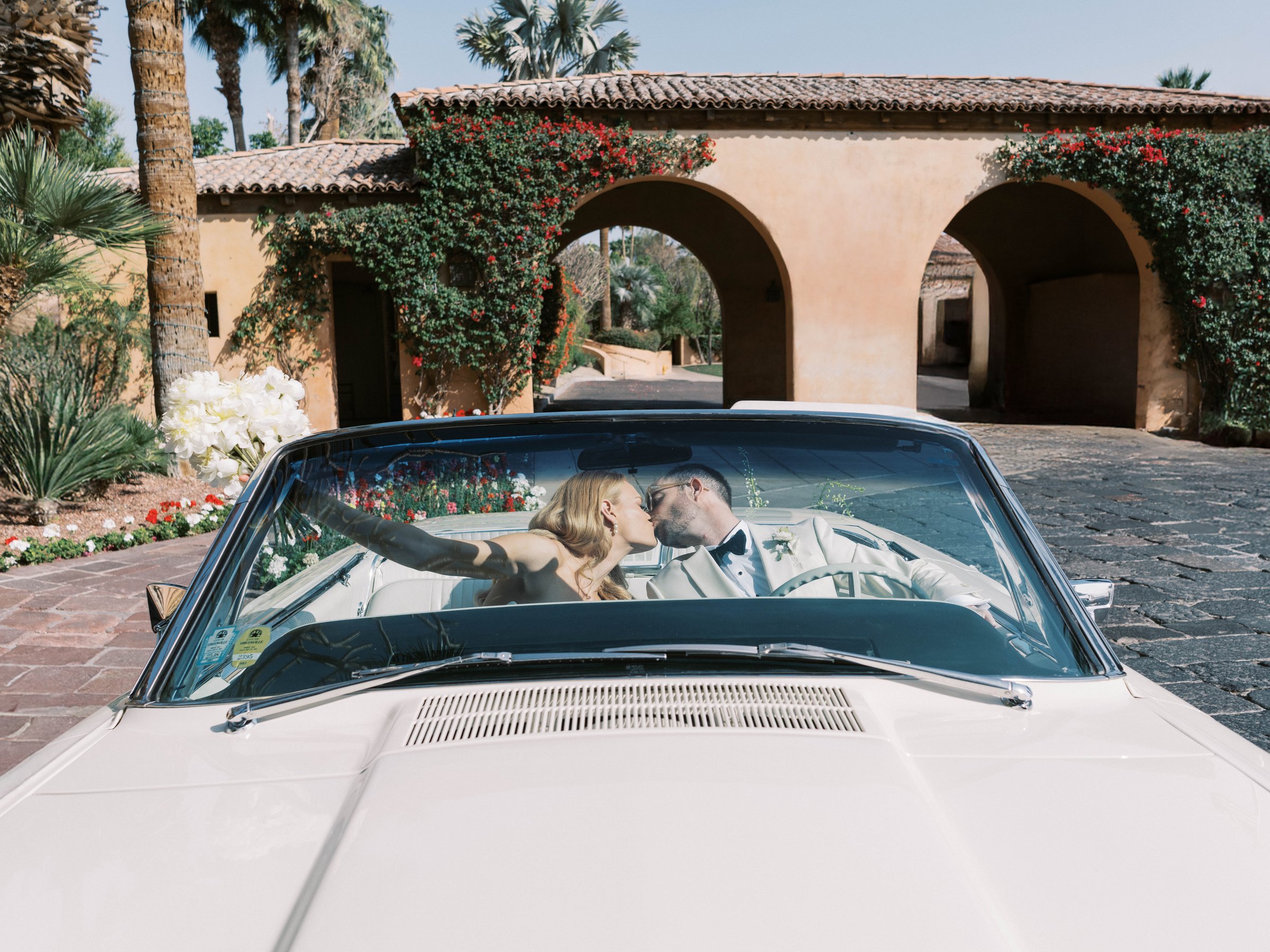 Meg and Peter kiss in a white vintage convertible beneath the Royal Palms arches