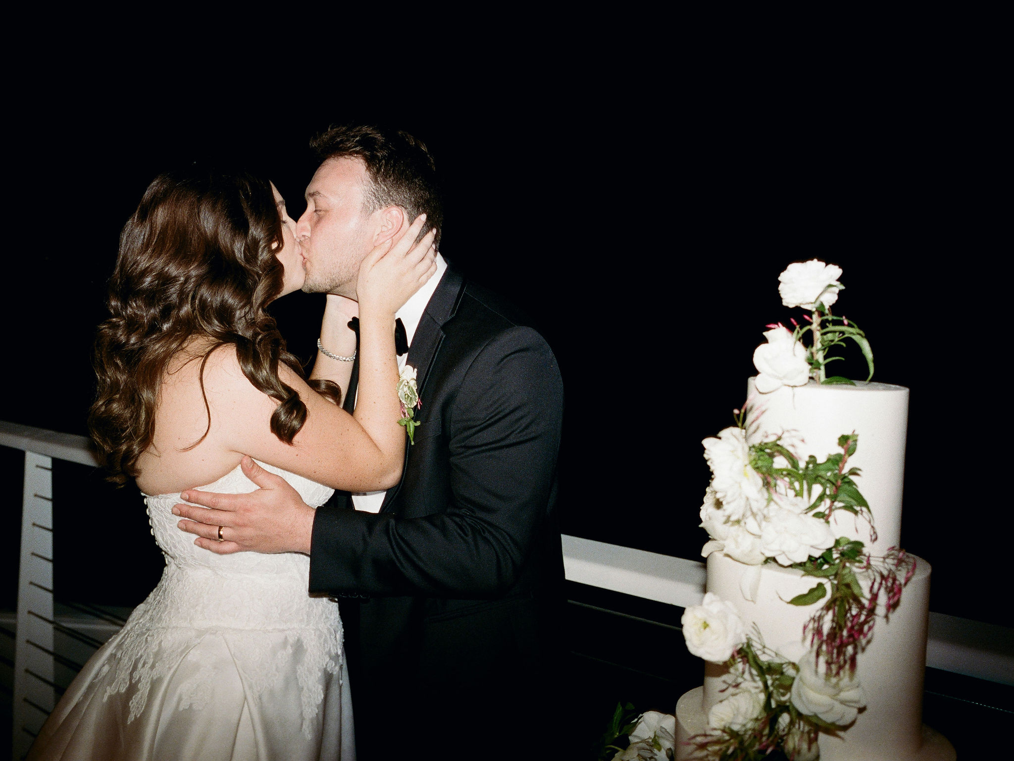 Taylor and Ryan kissing beside their flower-covered wedding cake on film.