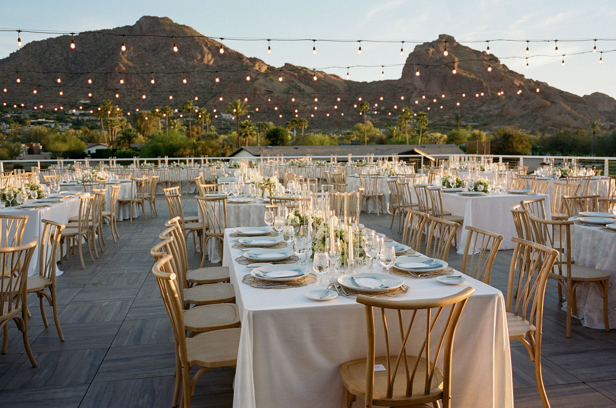 Outdoor reception tables beneath string lights with Camelback Mountain beyond.
