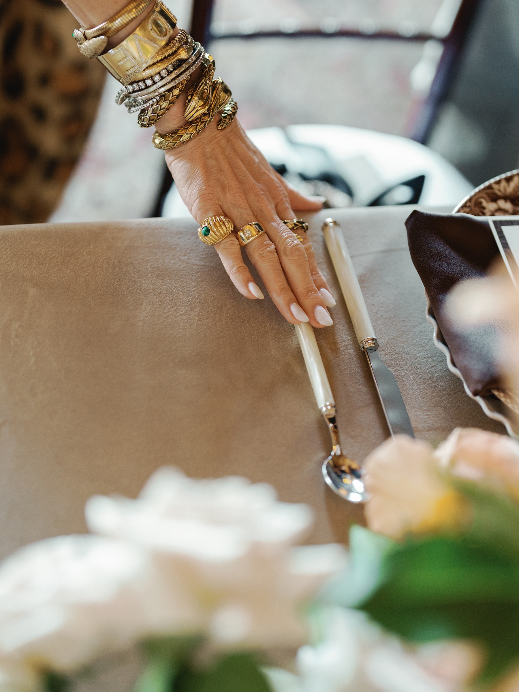 Hand with stacked gold bracelets and rings adjusting a place setting beside white roses