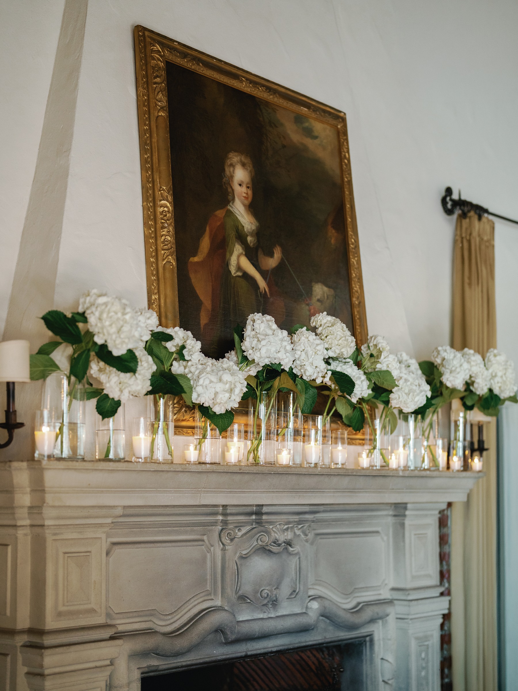 Stone fireplace mantel lined with white hydrangea and candles beneath an oil portrait