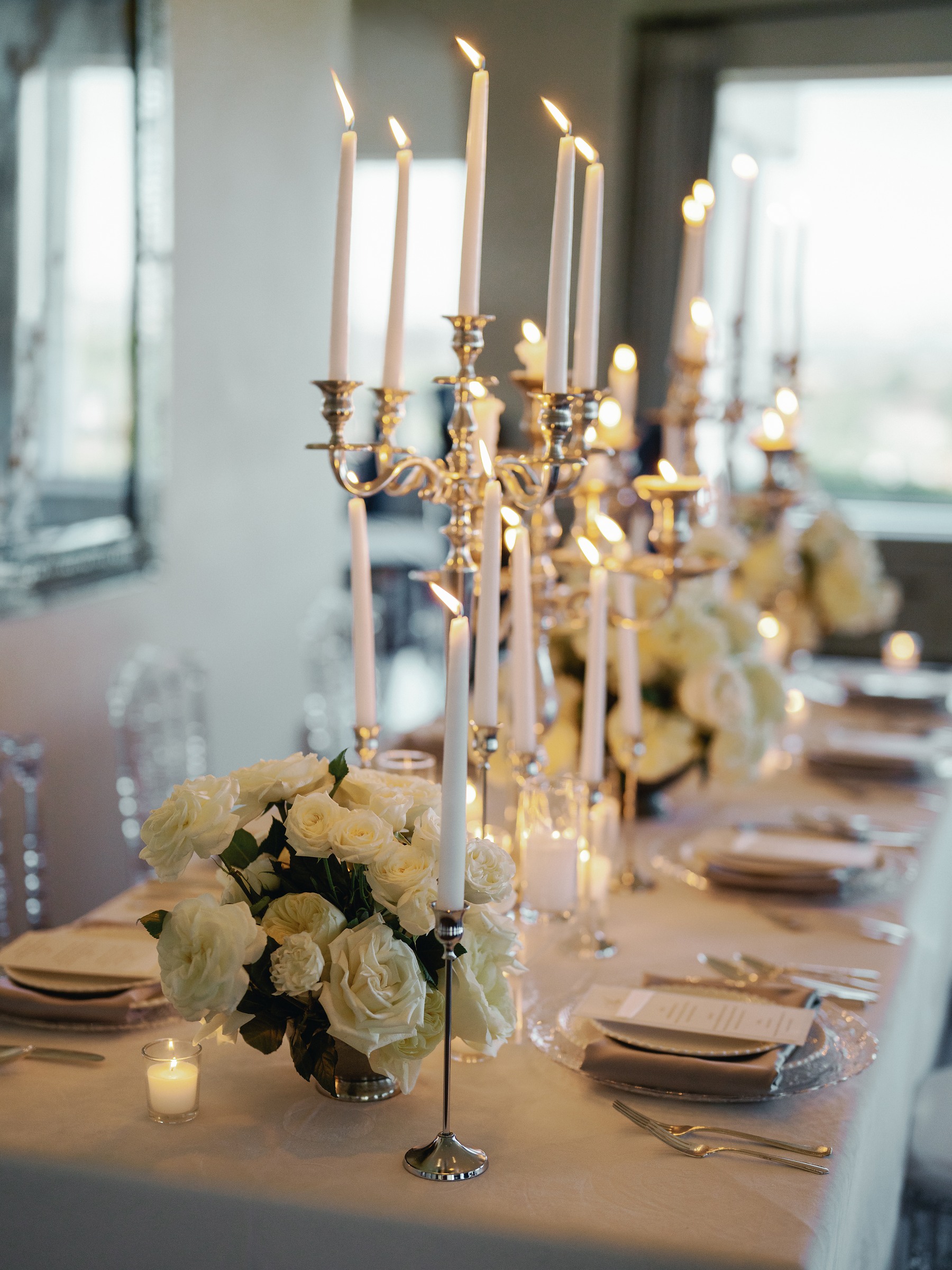 Silver candelabras and white roses down a candlelit table with clear ghost chairs